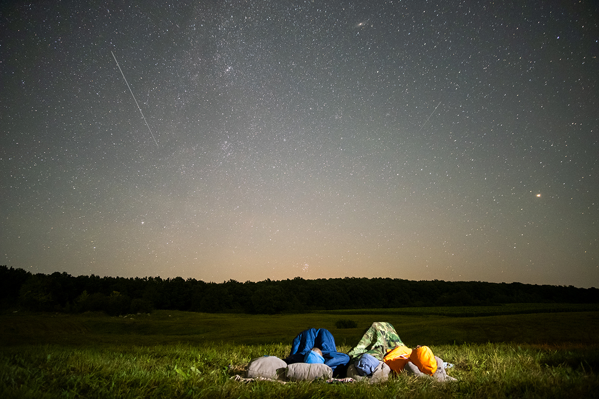The Perseid Meteor Shower Near Yellowstone: Your Complete 2026 Guide 4 People lying in sleeping bags in an open field watching a sky full of stars and Perseid Meteor Shower Near Yellowstone